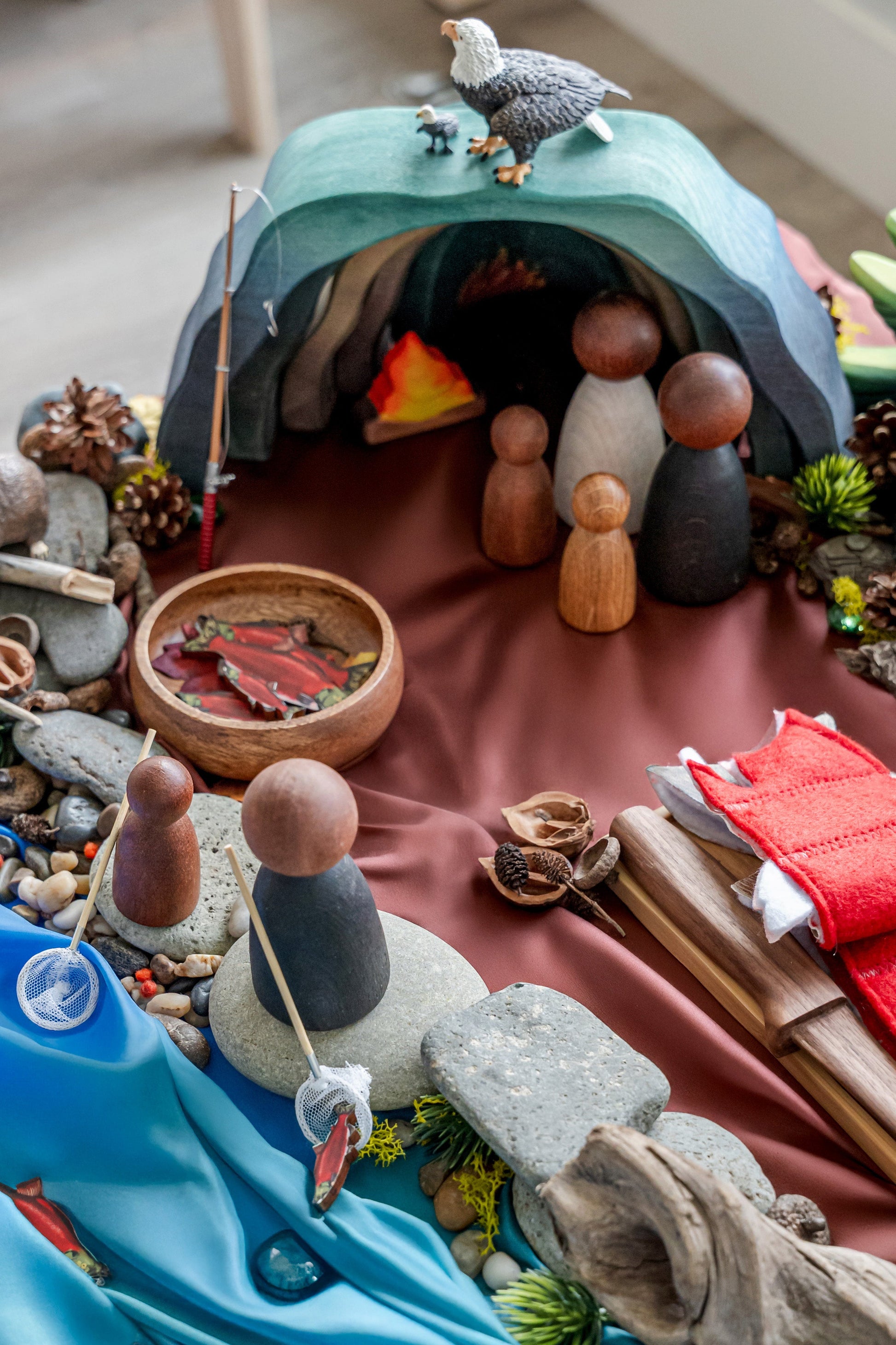 Children’s invitation to play with wooden figures, cave shelter, river fabric, and salmon storytelling setup for Indigenous-inspired learning
